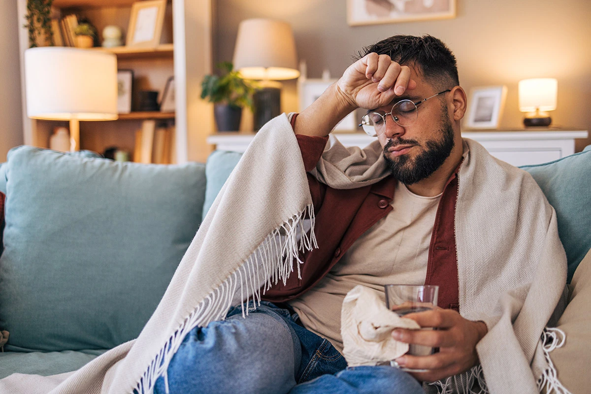 Man sitting on his couch with a blanket around him, holding the back of his hand up to his forehead